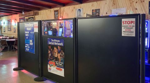 Matte Black tall hinged panel barriers enclose a video gaming area in a bar. Solid black panels screen the machines, and a custom panel with clear acrylic in its top quarter lets staff see inside.