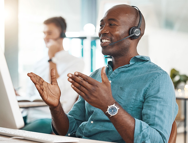 A Helpdesk support professional smiling and talking into his headset, smiling at his monitor as he resolves a customer's issue over a phone call.